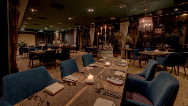 Een ruime eetzaal met meerdere tafels gedekt, met kaarsen en helder glaswerk.