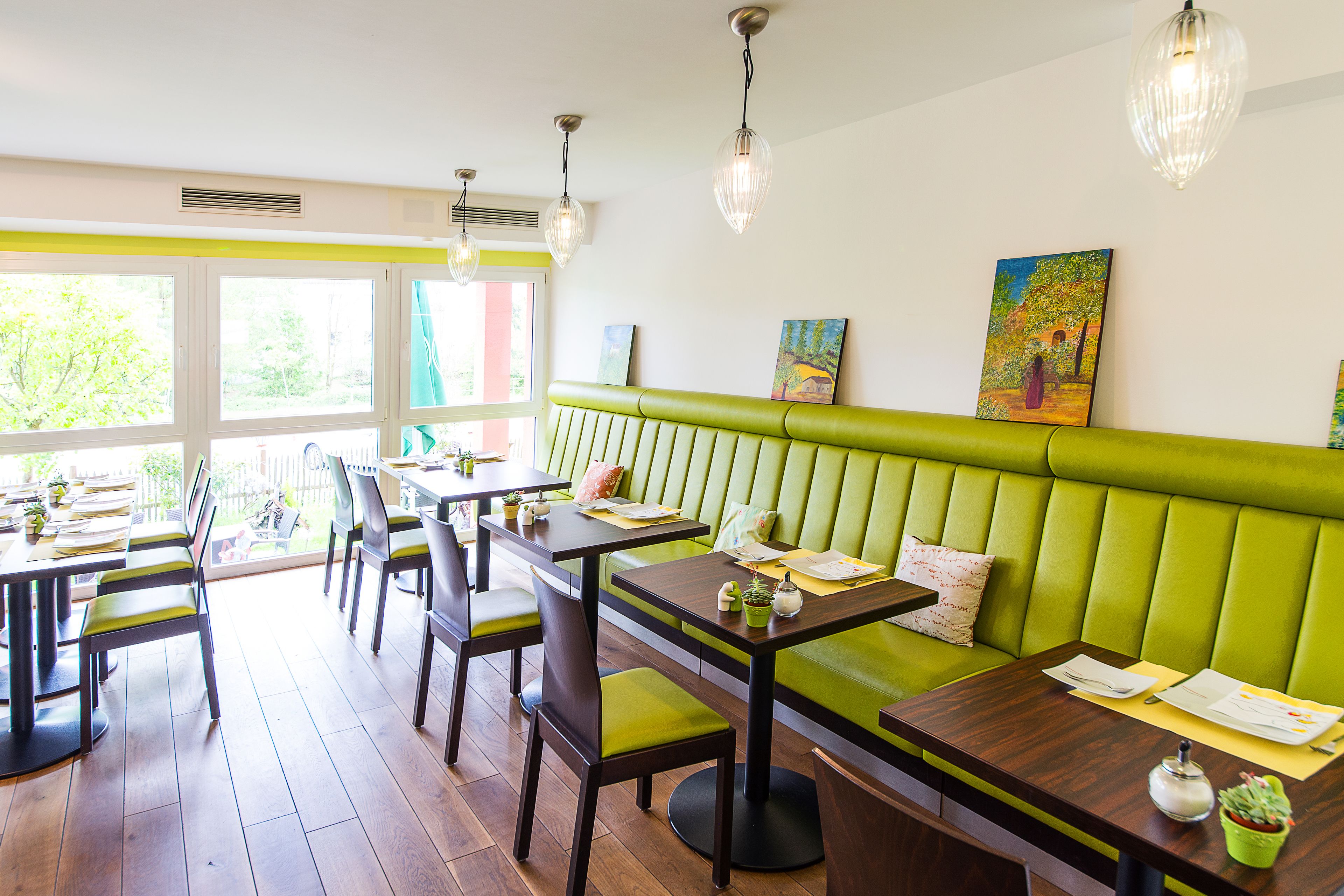 Ein Restaurant mit grünen Sitzbänken, Holztischen und -stühlen. Hängende Lampen und Bilder schmücken die Wände.