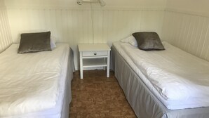 Una habitación con dos camas individuales, cada una con una almohada y una mesita de noche. El suelo es de madera y las paredes son blancas.