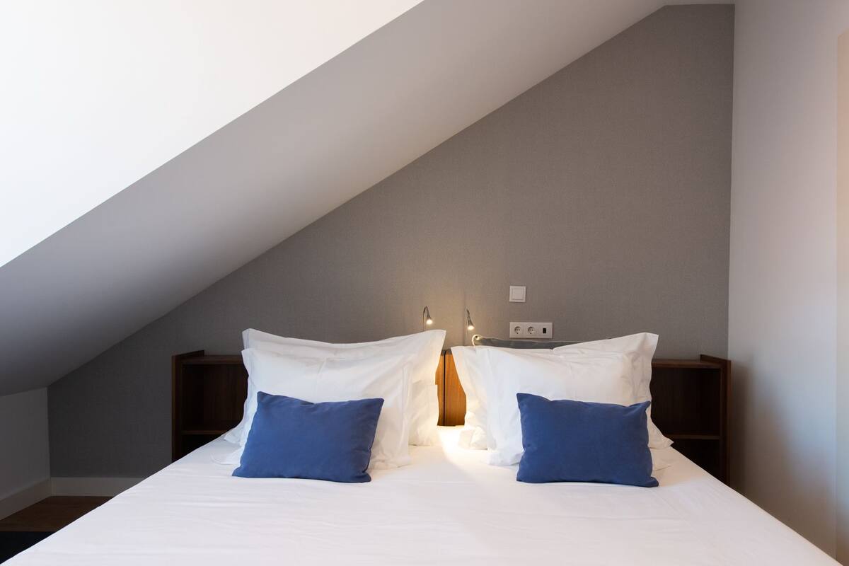 A bedroom with a sloped ceiling, a bed with white and blue bedding, two bedside tables, and wall-mounted reading lights.