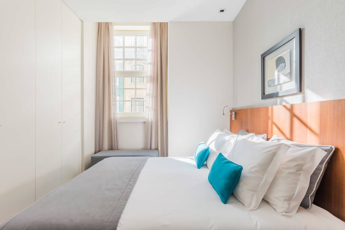 A hotel room with a bed, a window with curtains, a framed picture on the wall, and a wooden headboard.