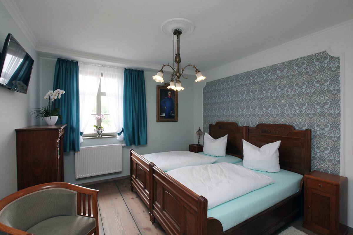 Ein Schlafzimmer mit zwei hölzernen Betten, einem Nachttisch aus Holz, einem Sessel, einem Fernseher und einem Fenster mit Vorhängen.