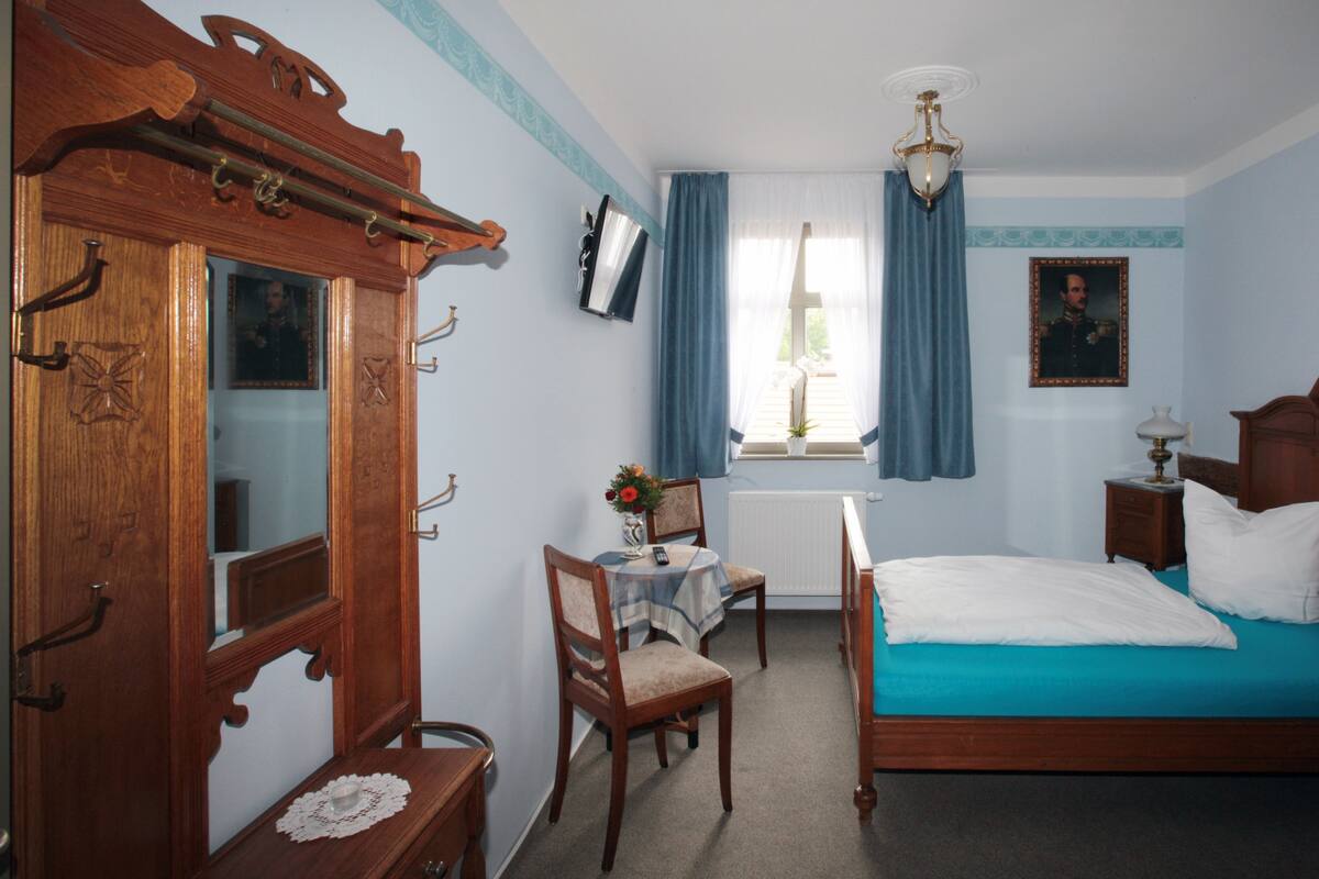 Ein Schlafzimmer mit einem hölzernen Bett, einem Schreibtisch mit Stuhl, einem Fenster mit Vorhängen und einem Heizkörper.