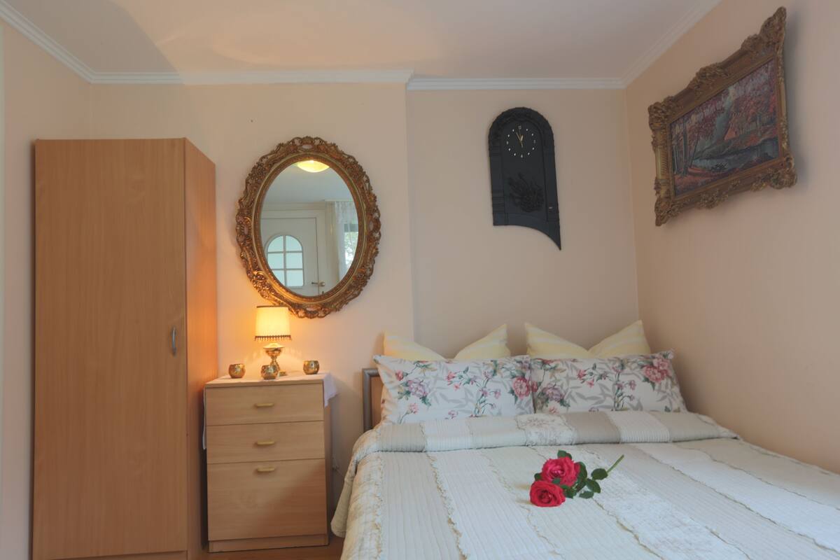 Une chambre à coucher avec une armoire en bois, une table de chevet avec une lampe, un lit avec une literie à motifs floraux, un miroir, une horloge et un tableau encadré accroché au mur.