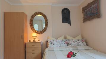 Une chambre à coucher avec une armoire en bois, une table de chevet avec une lampe, un lit avec une literie à motifs floraux, un miroir, une horloge et un tableau encadré accroché au mur.