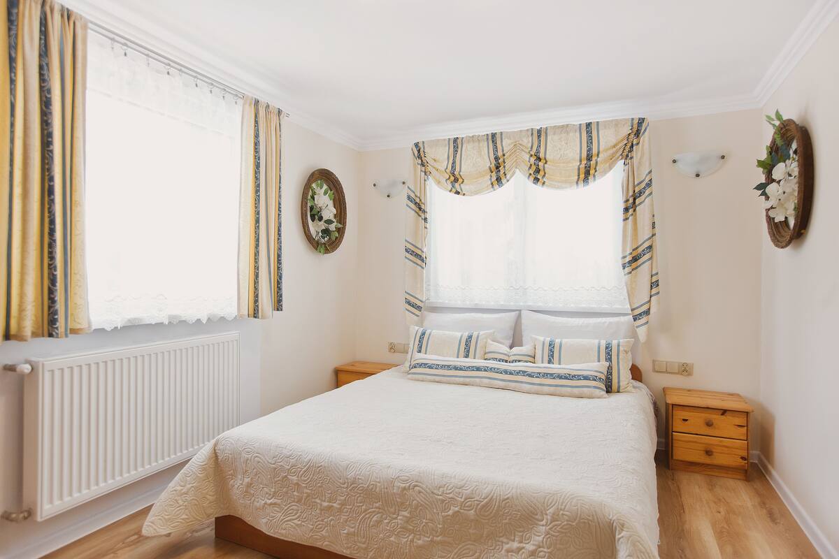 Une chambre à coucher avec un lit, des tables de chevet en bois, un radiateur et une fenêtre avec des rideaux.