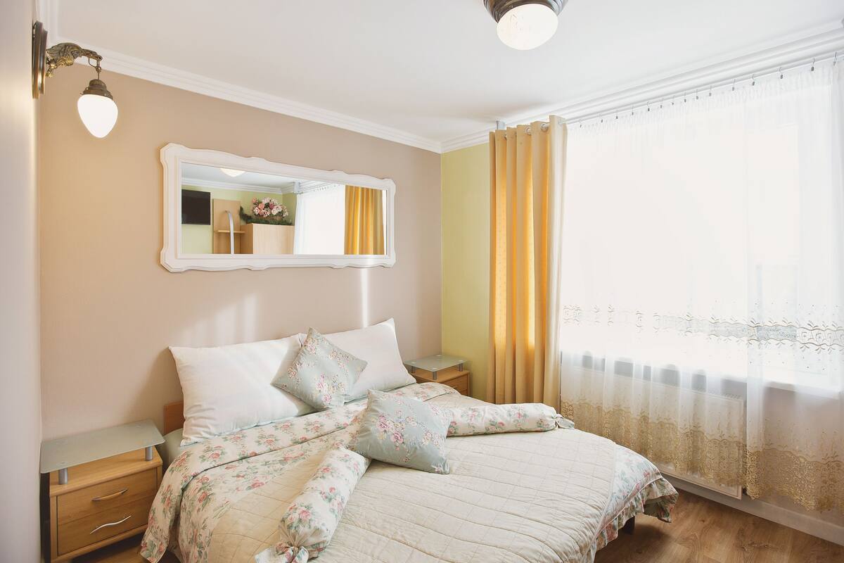 Une chambre à coucher avec un lit, une table de chevet, un miroir et une fenêtre avec des rideaux.