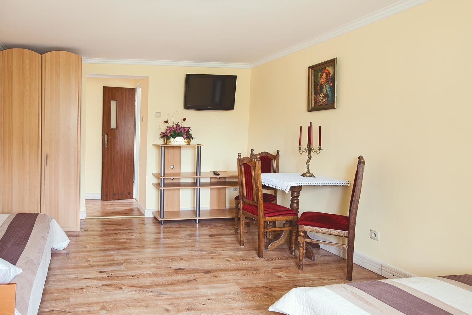 Una stanza con pavimento in legno, un letto con lenzuola a righe, un armadio in legno, un tavolo da pranzo con sedie, una televisione a parete e un quadro.