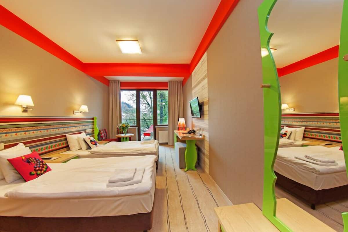 A hotel room with two beds, a desk, a chair, a TV, and a window with a view of trees.