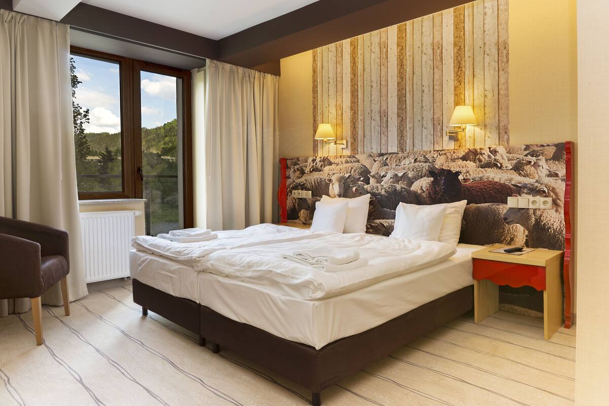 A hotel room with a large bed, two bedside tables, a chair, a window with curtains, and a wooden wall with a sheep painting.