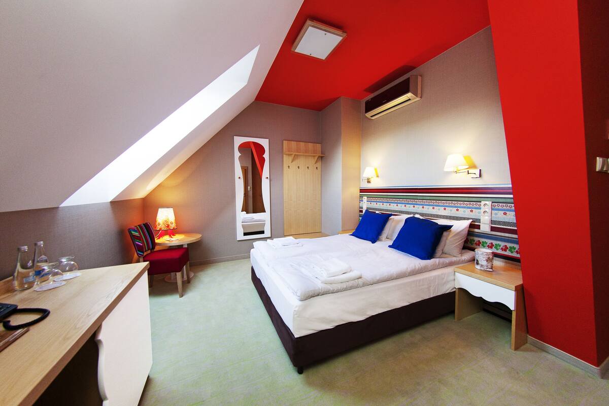 A hotel room with a bed, bedside table, desk, chair, and a small table with a lamp.