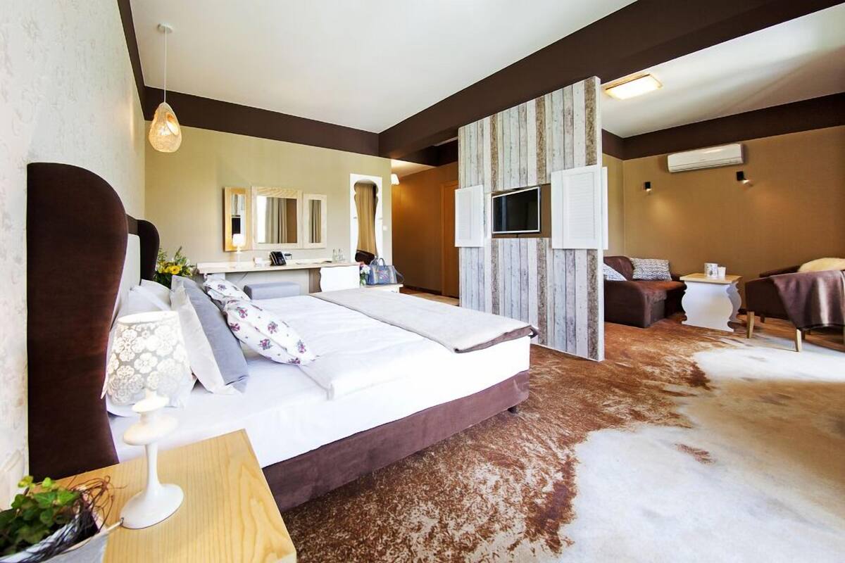 A modern hotel room with a large bed, a sitting area with a sofa and armchairs, a flat-screen TV mounted on the wall, and a large mirror above the bed.