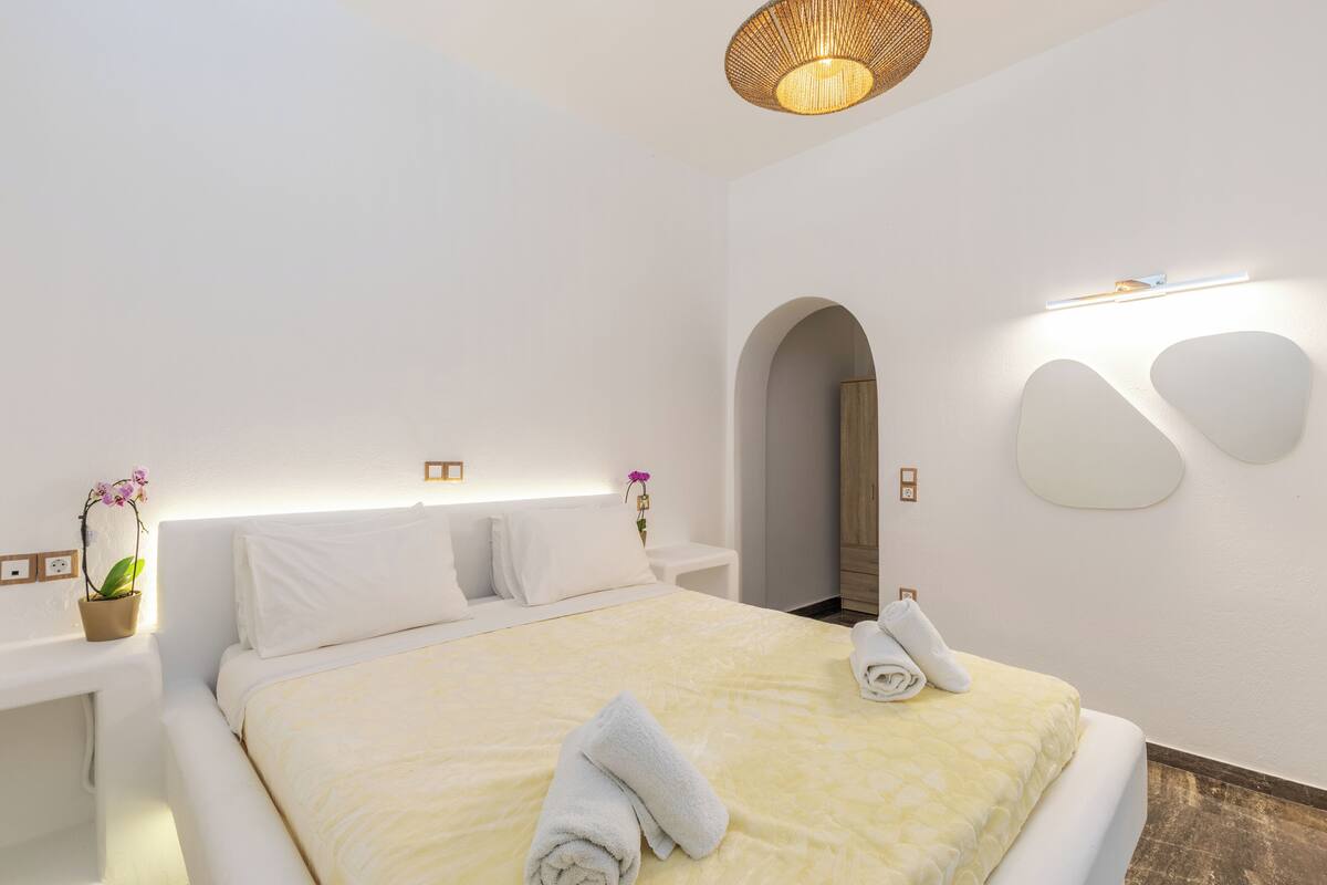 Une chambre d’hôtel moderne avec un grand lit, une table de chevet agrémentée d’une plante, un miroir et un luminaire fixé au mur.