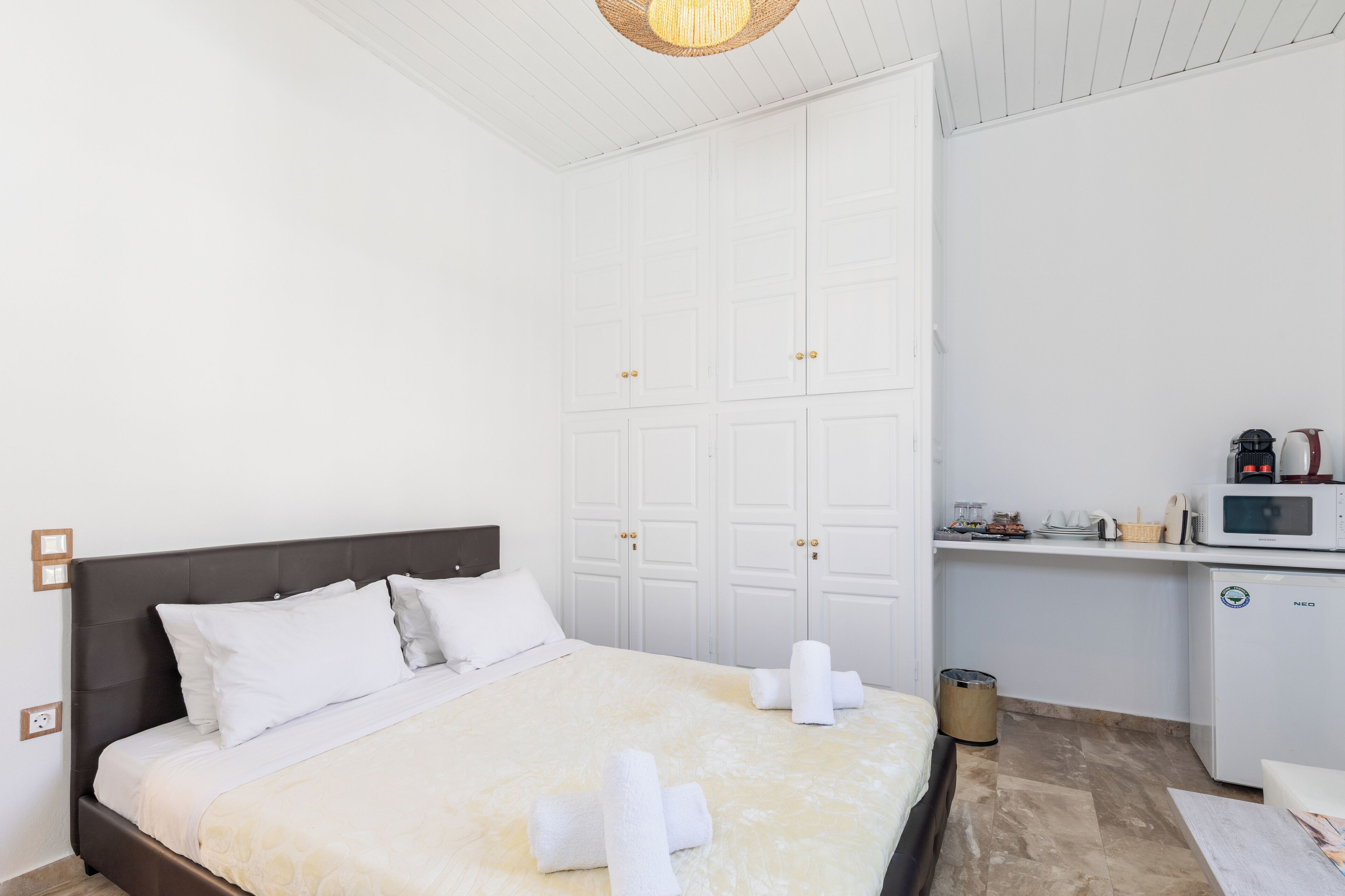 Une chambre d’hôtel avec un lit, une armoire blanche, une kitchenette et une poubelle.