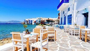 A seaside terrace with white and blue buildings, stone flooring, and outdoor seating. - Mykonian Breeze (Mykonos)