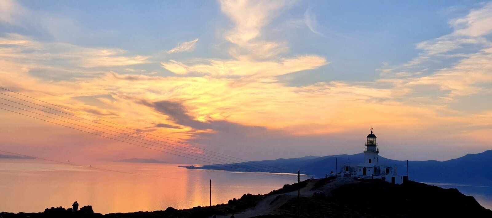 A coastal scene with a lighthouse on a hill overlooking a calm sea and distant mountains.