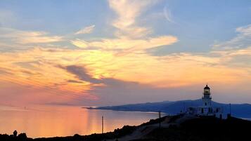 A coastal scene with a lighthouse on a hill overlooking a calm sea and distant mountains.