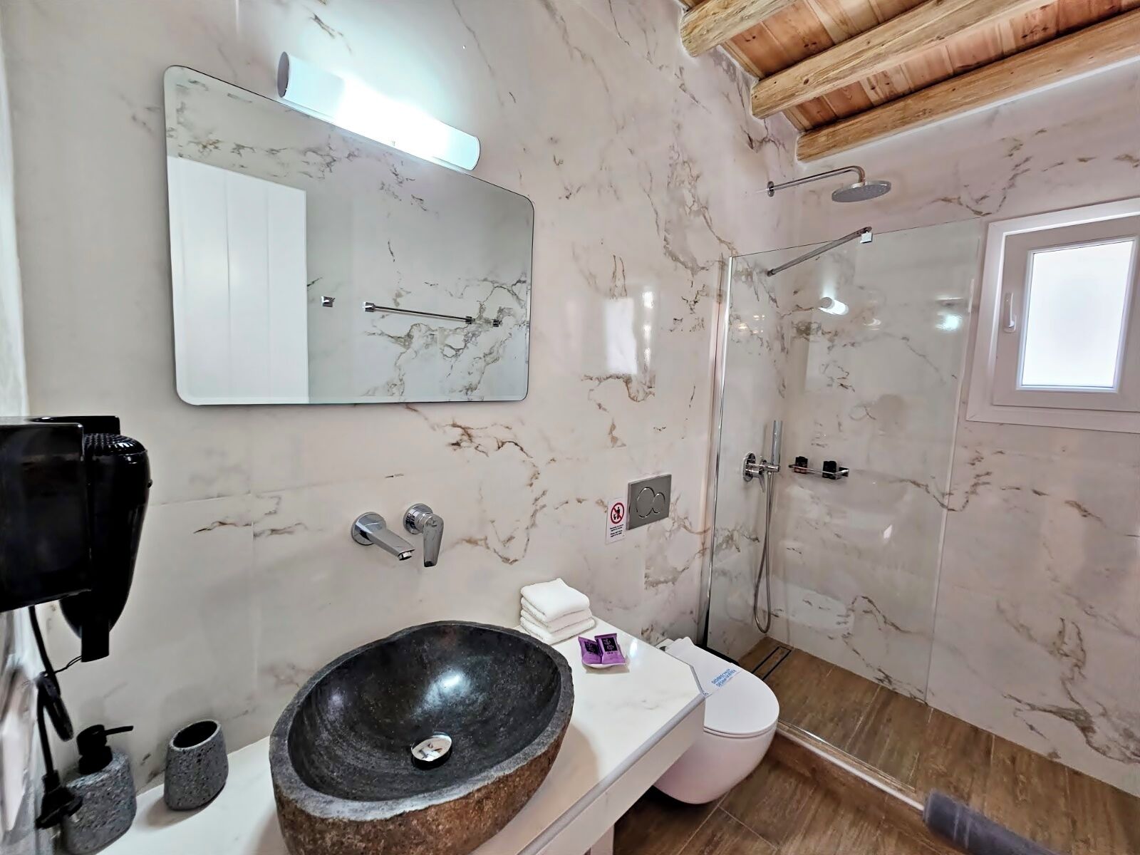 A bathroom with a marble wall, a large mirror, a black vessel sink, a toilet, and a shower area.