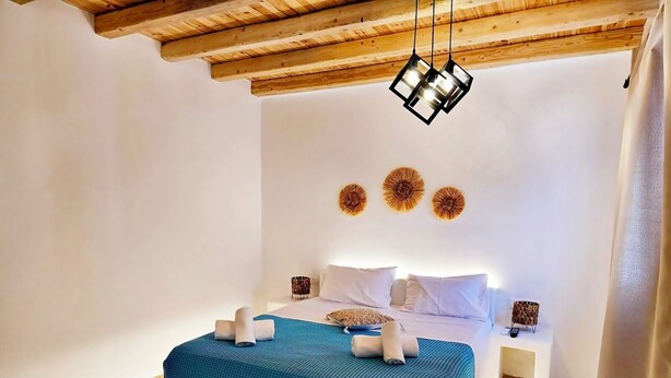A bedroom with a wooden ceiling, a bed with a blue bedspread, white pillows, and decorative wall art. - Mykonian Breeze (Mykonos)