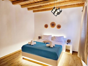 A bedroom with a wooden ceiling, a bed with a blue bedspread, white pillows, and decorative wall art. - Mykonian Breeze (Mykonos)