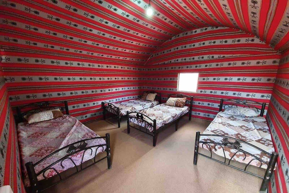 A room with four beds arranged in a corner, each with a patterned bedspread and a pillow. The walls are adorned with a red and white striped pattern.