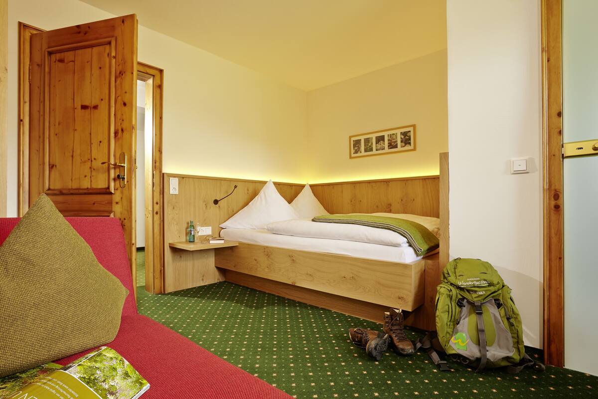 A wooden bed with white bedding, a green backpack, and a pair of boots on a green patterned carpet.