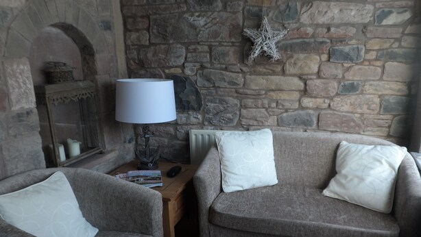 A cozy living room with two grey armchairs, a wooden side table, a lamp, a magazine, and a stone wall with a decorative star. - Midtown Farm Bed & Breakfast (Wigton)