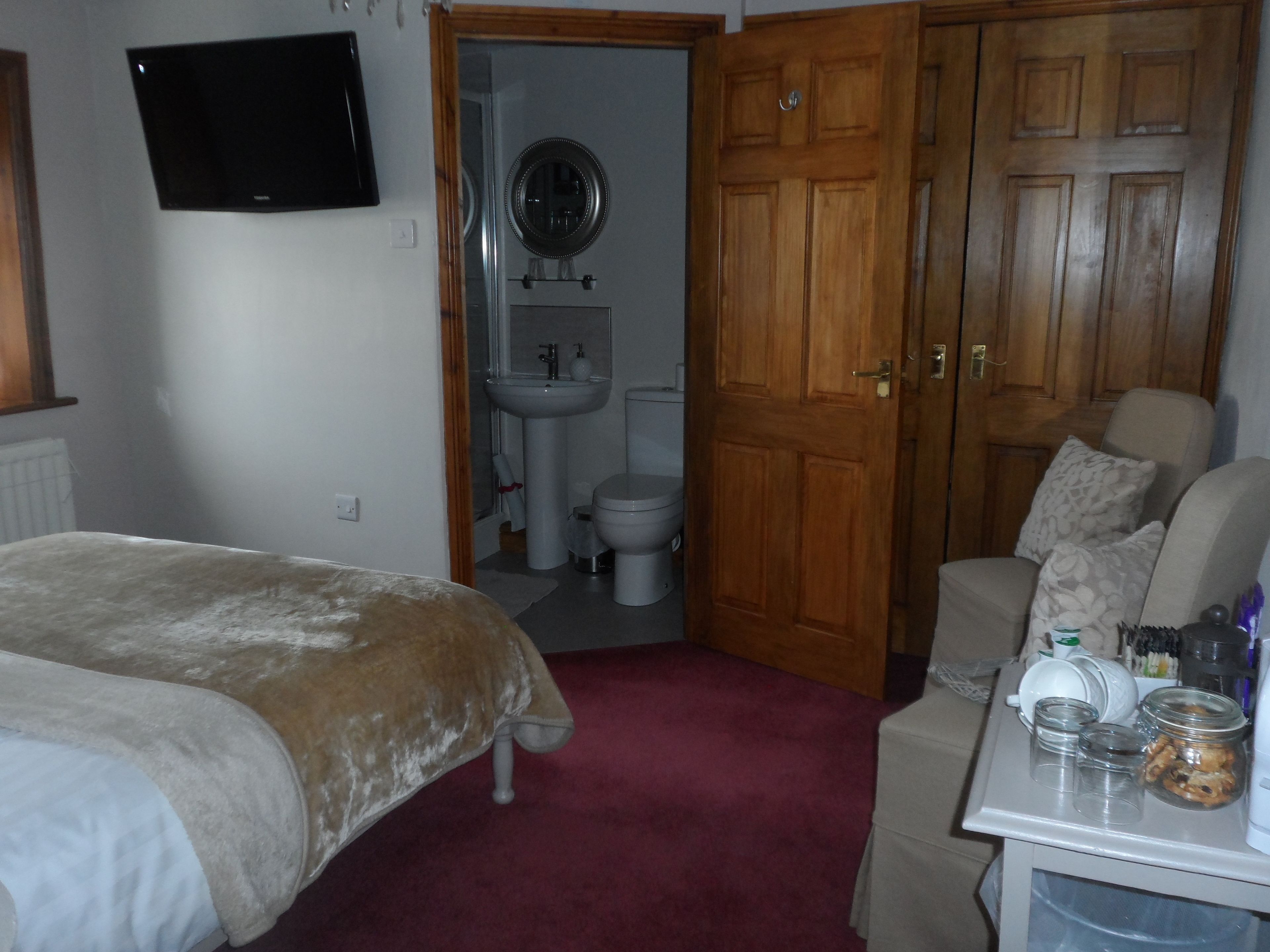 Un dormitorio con cama, un armario de madera, una mesita con vasos y un tarro, y un baño visible a través de una puerta abierta.