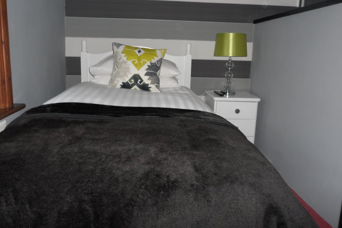 A bed with a black blanket, a pillow with a geometric pattern, and a bedside table with a lamp.