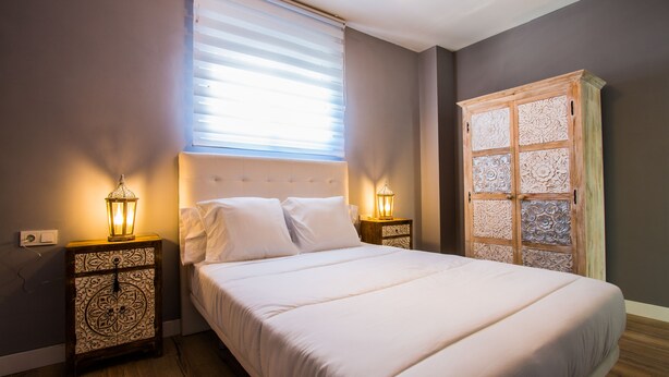 Una cama bien hecha con sábanas blancas, dos mesitas de noche con lámparas decorativas, una ventana con persianas y un armario de madera con paneles ornamentales.