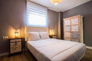 Una cama bien hecha con sábanas blancas, dos mesitas de noche con lámparas decorativas, una ventana con persianas y un armario de madera con paneles ornamentales.