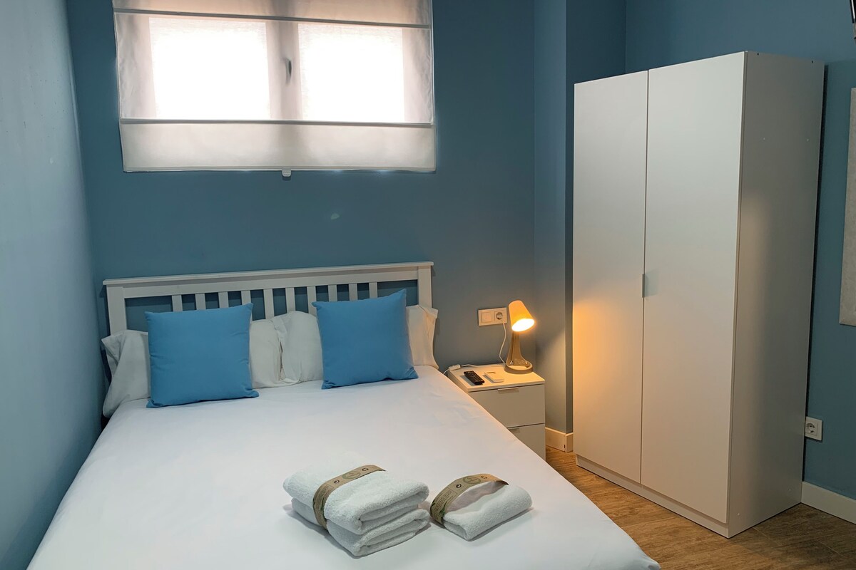 En pænt redt seng med blå og hvide puder, en sengetlampe og en stak håndklæder.