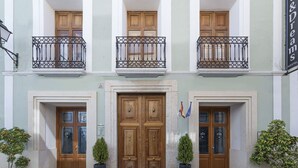Un edificio con tres puertas de madera, cada una con un pequeño balcón encima. Hay dos macetas con plantas a cada lado de la entrada.