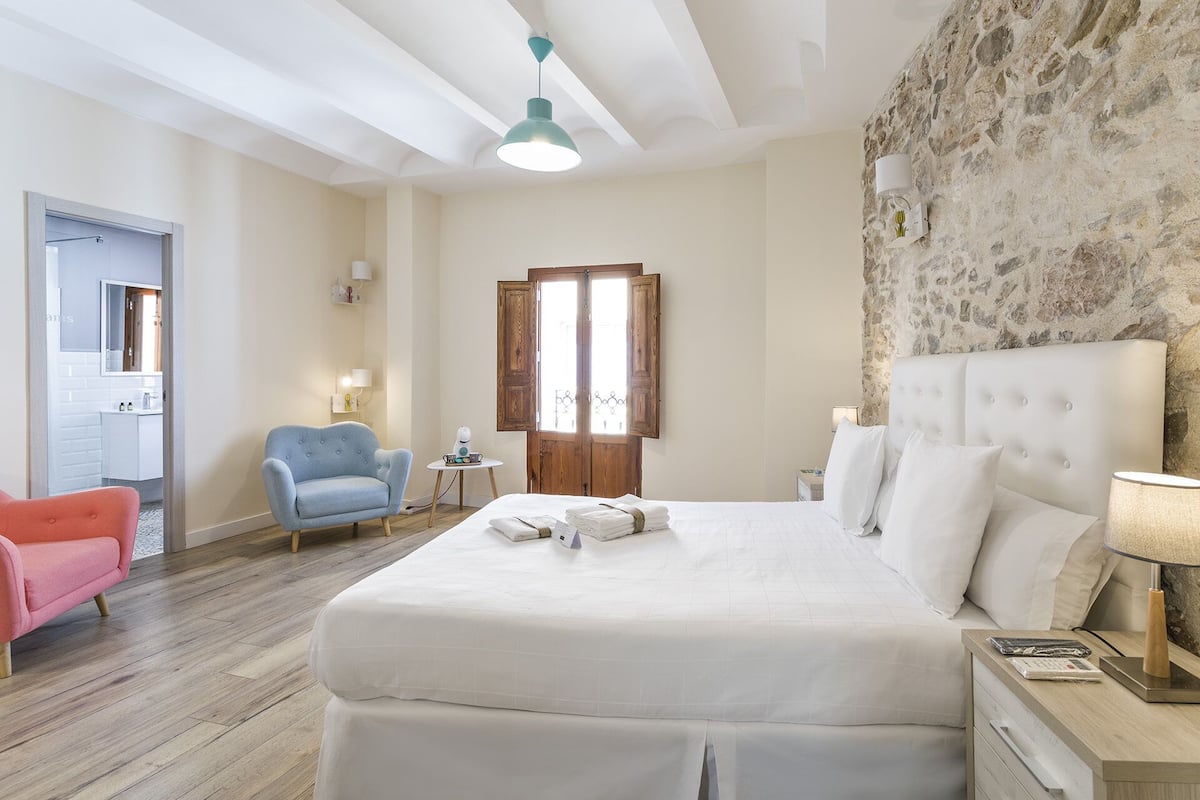 A bedroom with a large bed, a wooden door, a stone wall, a blue chair, and a pink chair.