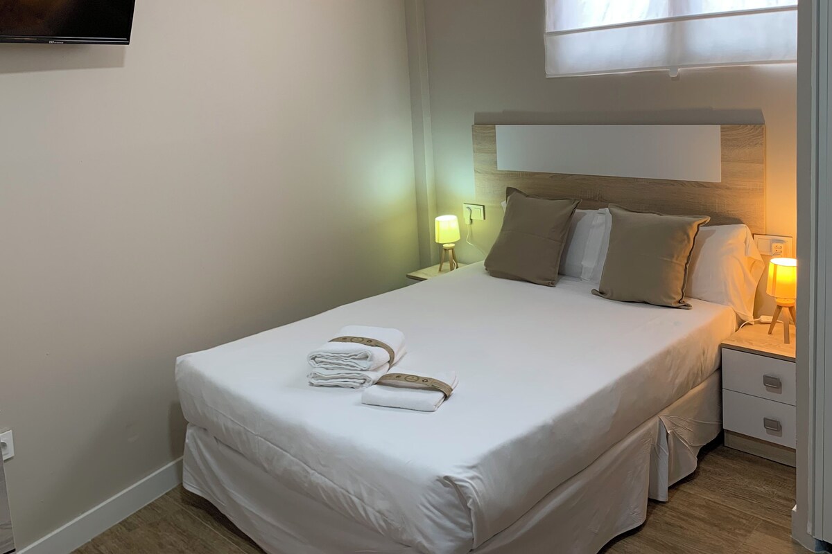 A neatly made bed with white linens and a set of towels, a bedside lamp, and a television mounted on the wall.