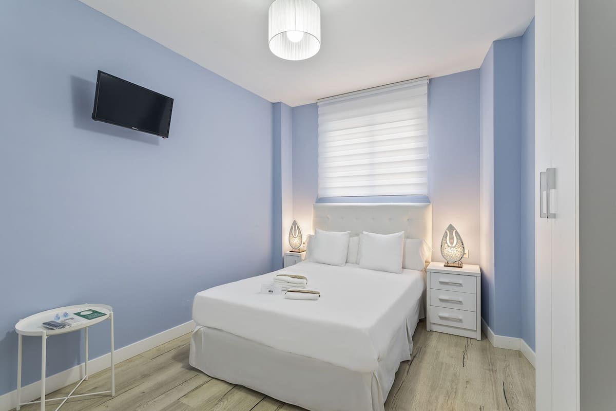 A bedroom with a bed, bedside tables, a TV, and a window with blinds.