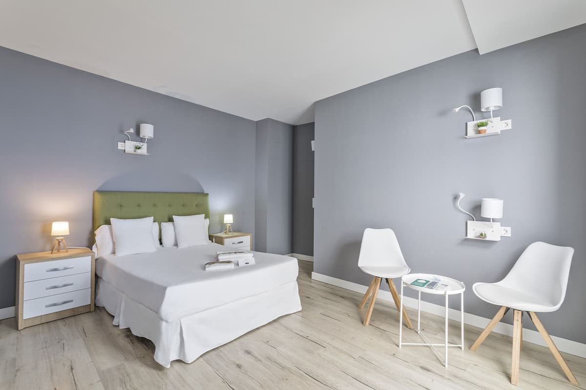 A modern hotel room with a large bed, bedside tables, a small table, two chairs, and wall-mounted lamps.