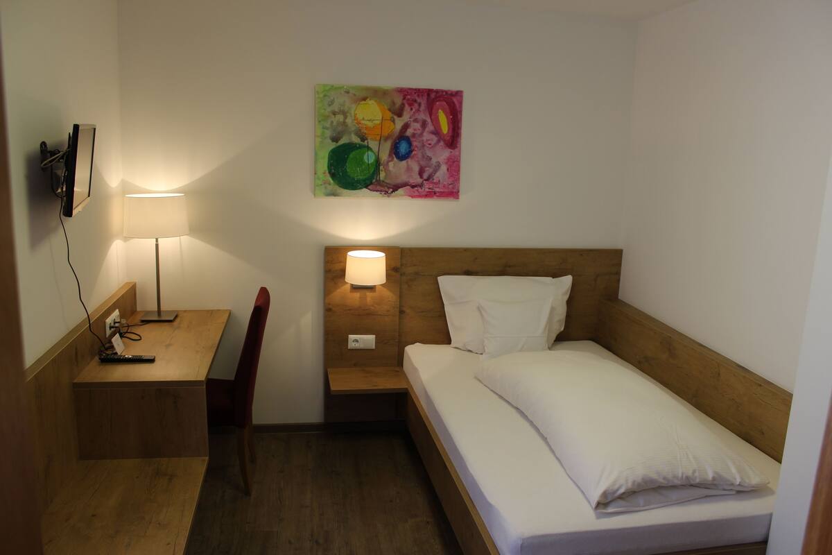 Une petite chambre d’hôtel, bien rangée, avec un bureau en bois, une chaise, une table de chevet avec une lampe, un lit avec du linge de lit blanc et un tableau abstrait coloré accroché au mur.