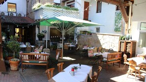Een terras buiten met tafels, stoelen, een groene parasol en potplanten.