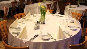 Een restaurant met ronde tafels gedekt voor diners, elk met witte tafelkleden, stoelen en een vaas met gele bloemen.