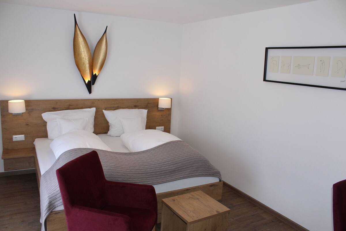Une chambre d’hôtel moderne avec une tête de lit en bois, un grand lit recouvert de draps blancs, une chaise rouge et une petite table de chevet en bois.