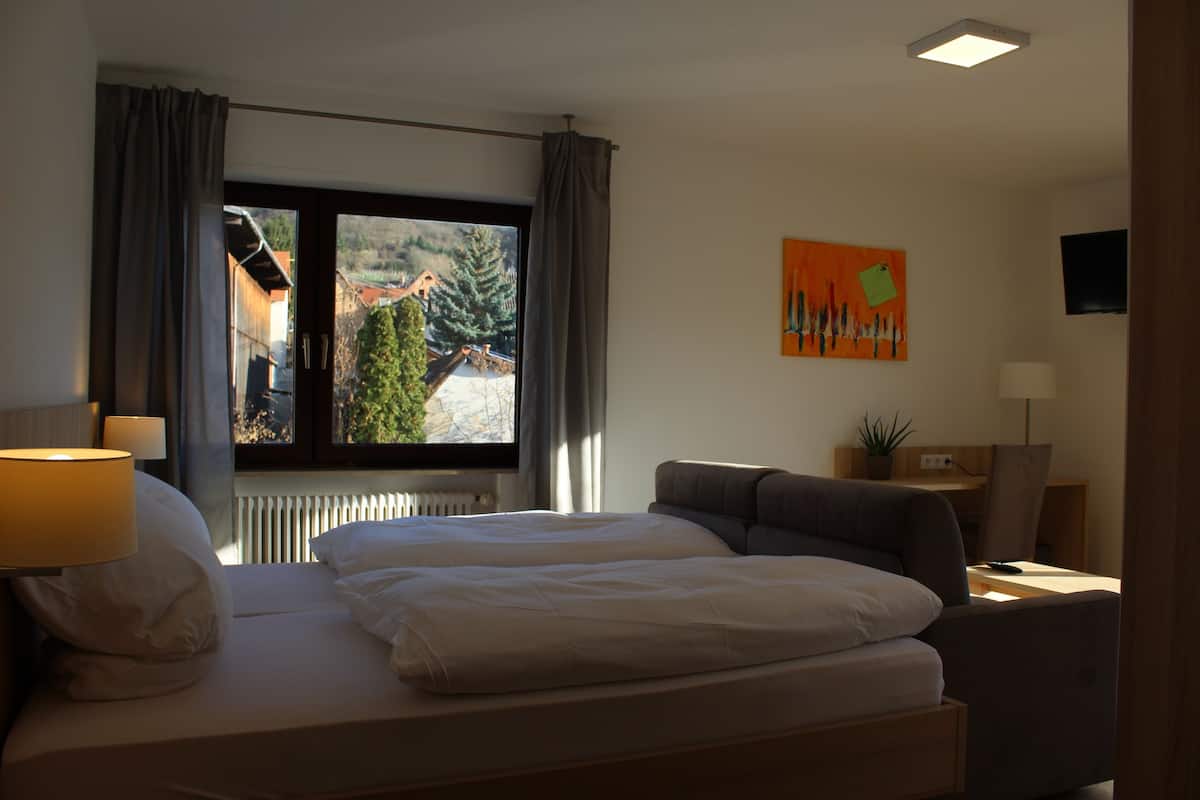 Une chambre d’hôtel comprenant un lit, un bureau, une chaise, une lampe et une fenêtre donnant sur des immeubles et des arbres.