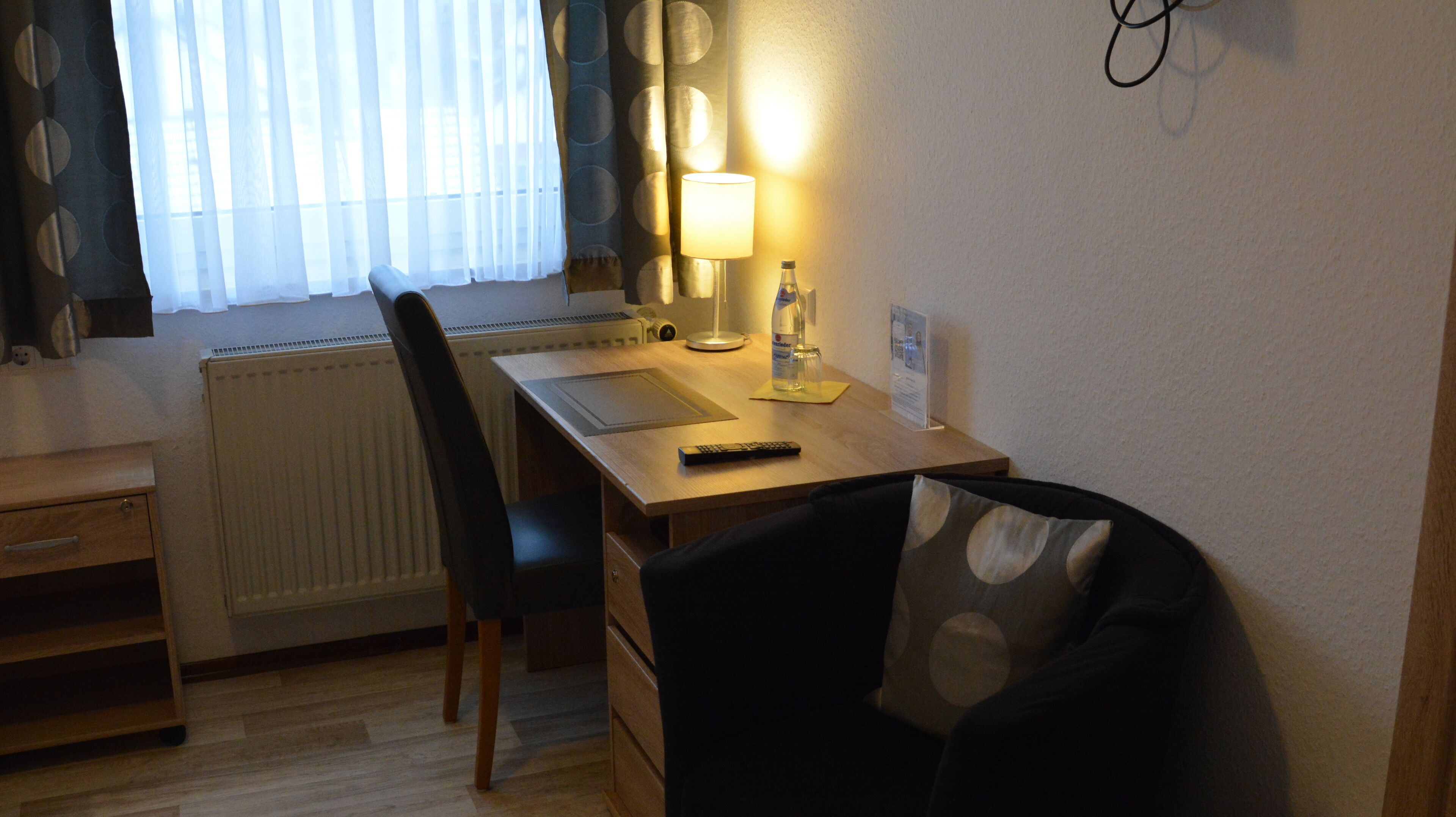Ein Hotelzimmer mit einem Holztisch, einem Stuhl, einer Lampe, einer Wasserflasche und einer Fernbedienung.