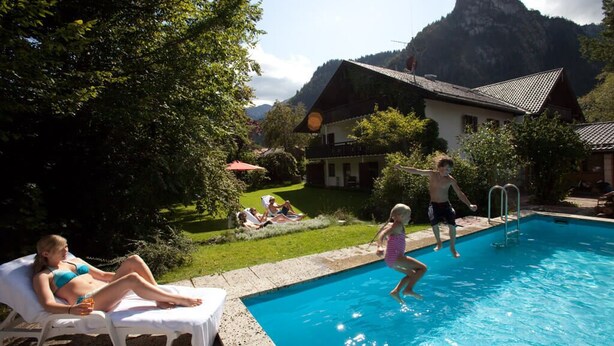 A swimming pool with people around it, a house, and mountains in the background. - DAS Posch Hotel (Oberammergau)
