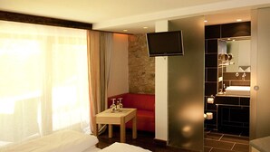 A hotel room with a bed, a small table with a bottle and glasses, a red sofa, a TV, and a bathroom with a sink and mirror. - DAS Posch Hotel (Oberammergau)