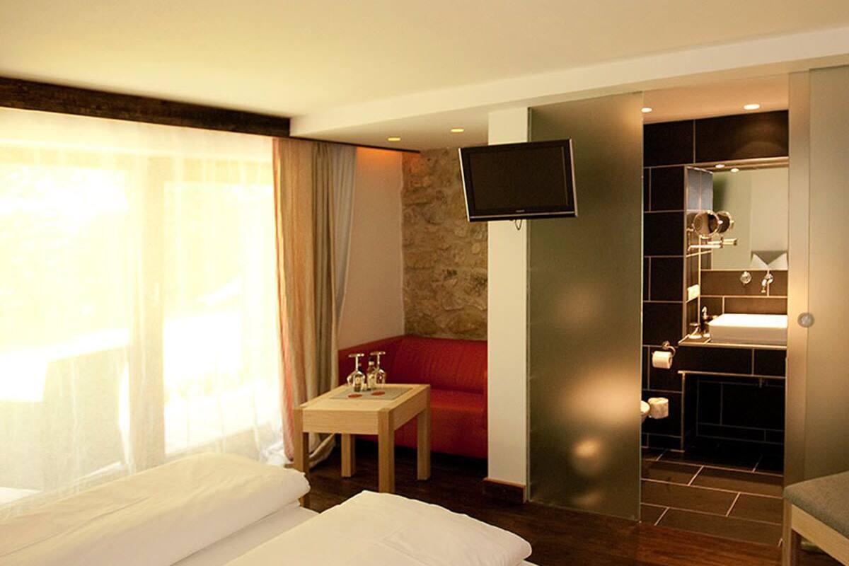 Una habitación de hotel con una cama, una mesita con una botella y vasos, un sofá rojo, un televisor y un baño con lavabo y espejo.