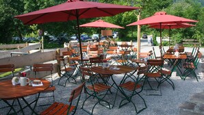 An outdoor cafe with multiple tables and chairs, red umbrellas, and a parking area in the background. - DAS Posch Hotel (Oberammergau)