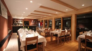 A restaurant with wooden tables and chairs, a bar area, and large windows. - DAS Posch Hotel (Oberammergau)