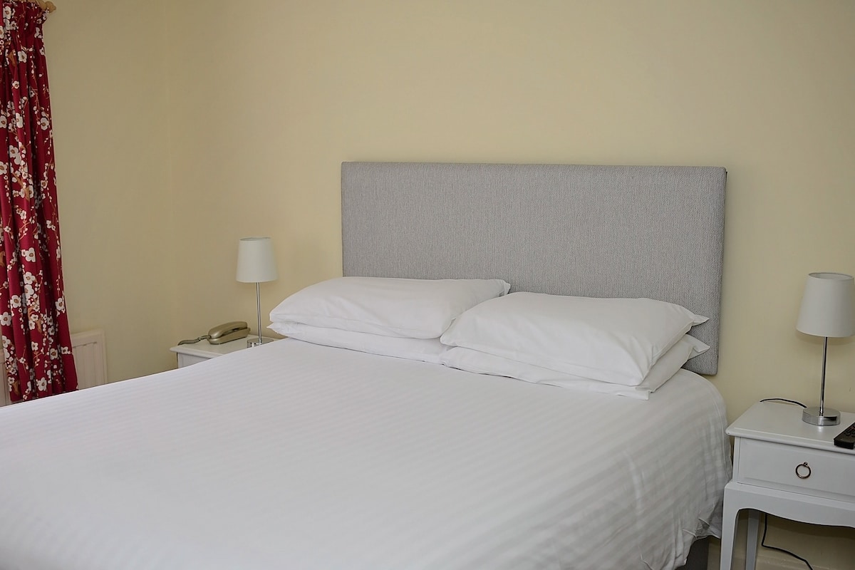A neatly made bed with white linens, a grey headboard, two bedside tables with lamps, and a red floral curtain.
