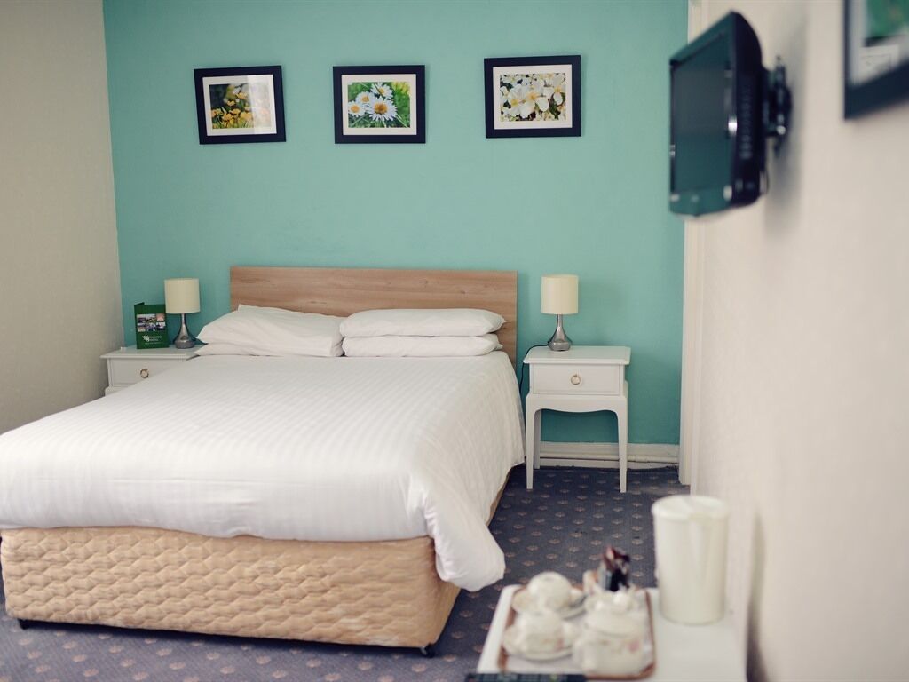 Una cama bien hecha con cabecera de madera, dos mesitas de noche con lámparas y tres cuadros florales enmarcados en la pared.